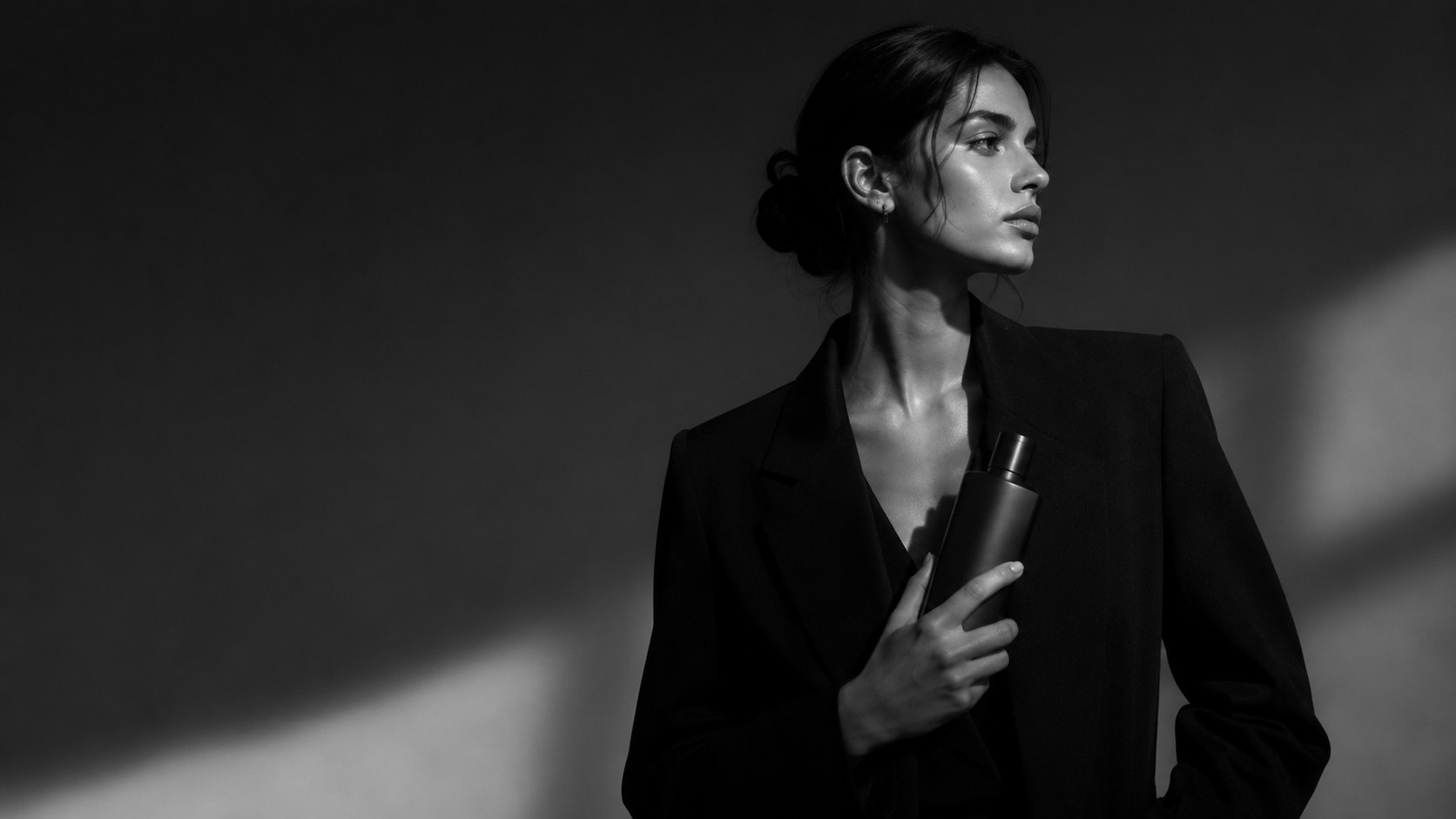 A model in a black blazer holds an unbranded bottle in soft window light, in black and white